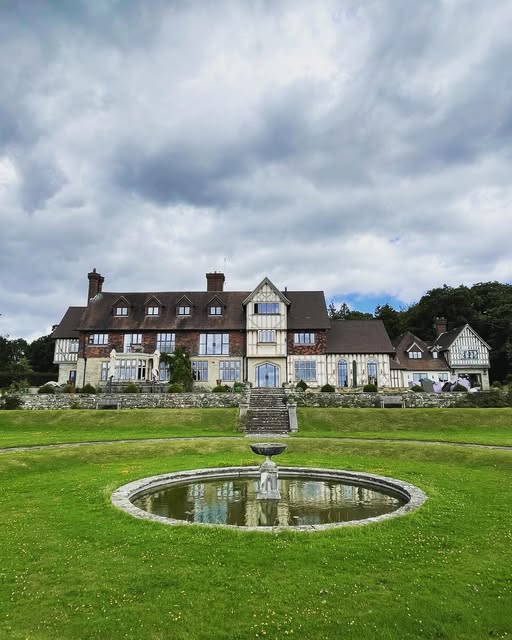 A large timber-framed country house with lawns and a circular fountain pond